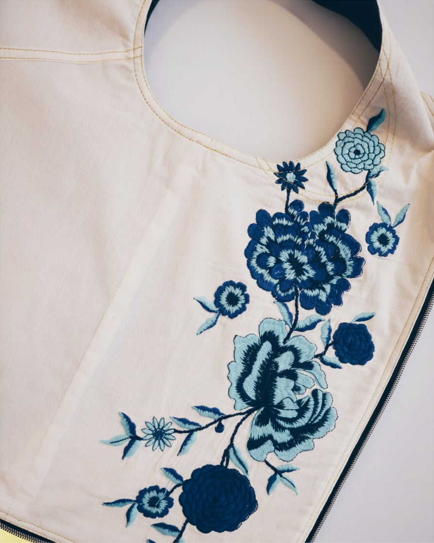 Hue Labs Floral Dreams vest bag in cream fabric with blue floral embroidery and a silver metal zipper detail.
